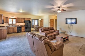 A spacious living room with a brown leather sofa, a dining area with a table and chairs, a kitchen with wooden cabinets and a refrigerator, and a ceiling fan.