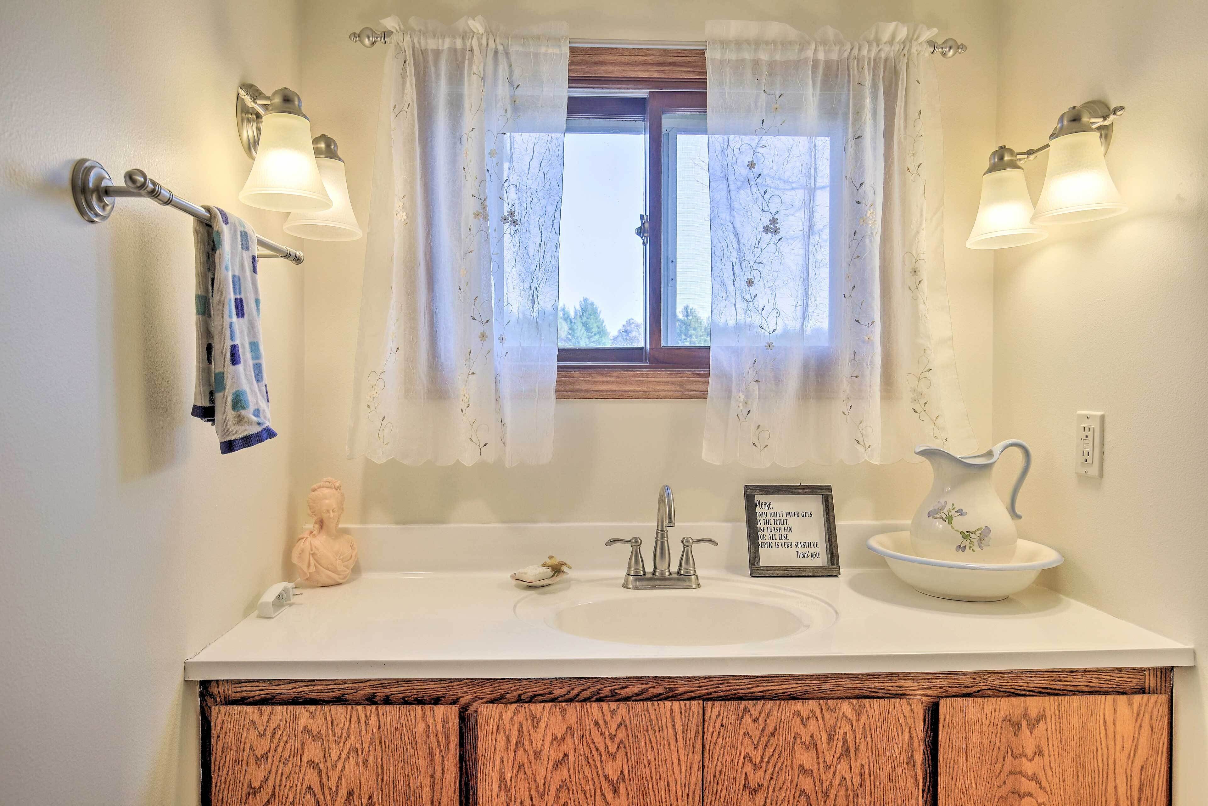 Une salle de bain avec un lavabo blanc, un miroir, une fenêtre ornée de rideaux en dentelle et un vase décoratif.