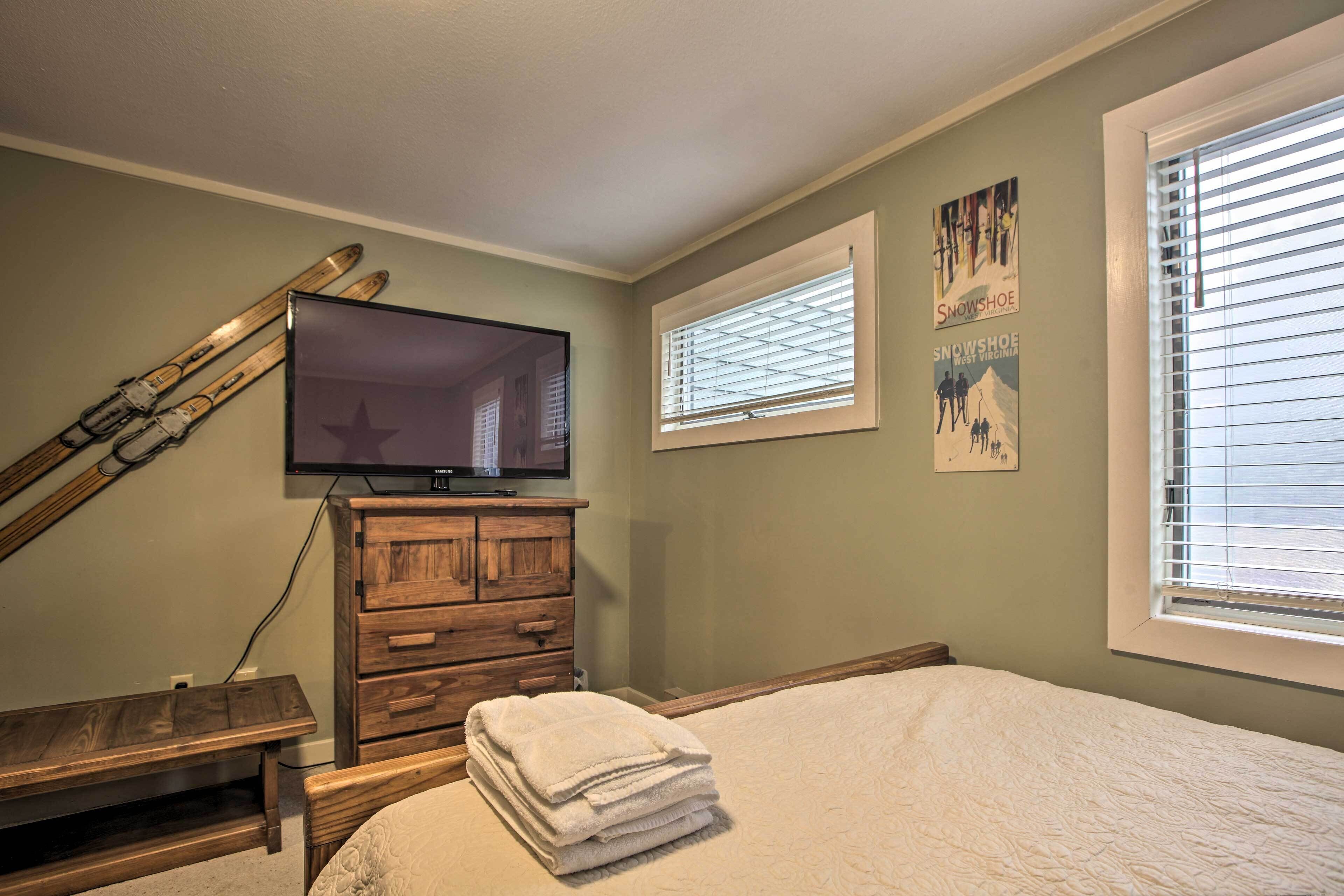Quarto com cama, cómoda de madeira, TV fixada na parede e janela com persianas.