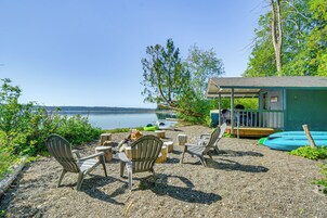 Un espacio al aire libre junto al lago con sillas Adirondack, un hogar para fuego y un pequeño edificio con porche.