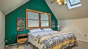 A bedroom with a sloped ceiling, a large window with blinds, a bed with a patterned blanket, and two wall-mounted lamps. - Peaceful Russellville Cabin Close to Lakes! (Russellville)