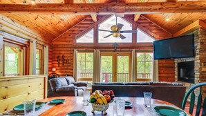 A wooden cabin interior with a dining table set for four, a fireplace, and a large TV mounted on the wall.