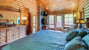 A log cabin interior with a large bed, wooden dresser, stone fireplace, and a television mounted on the wall.