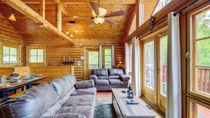 A log cabin interior with a large leather couch, a wooden dining table, and a ceiling fan.