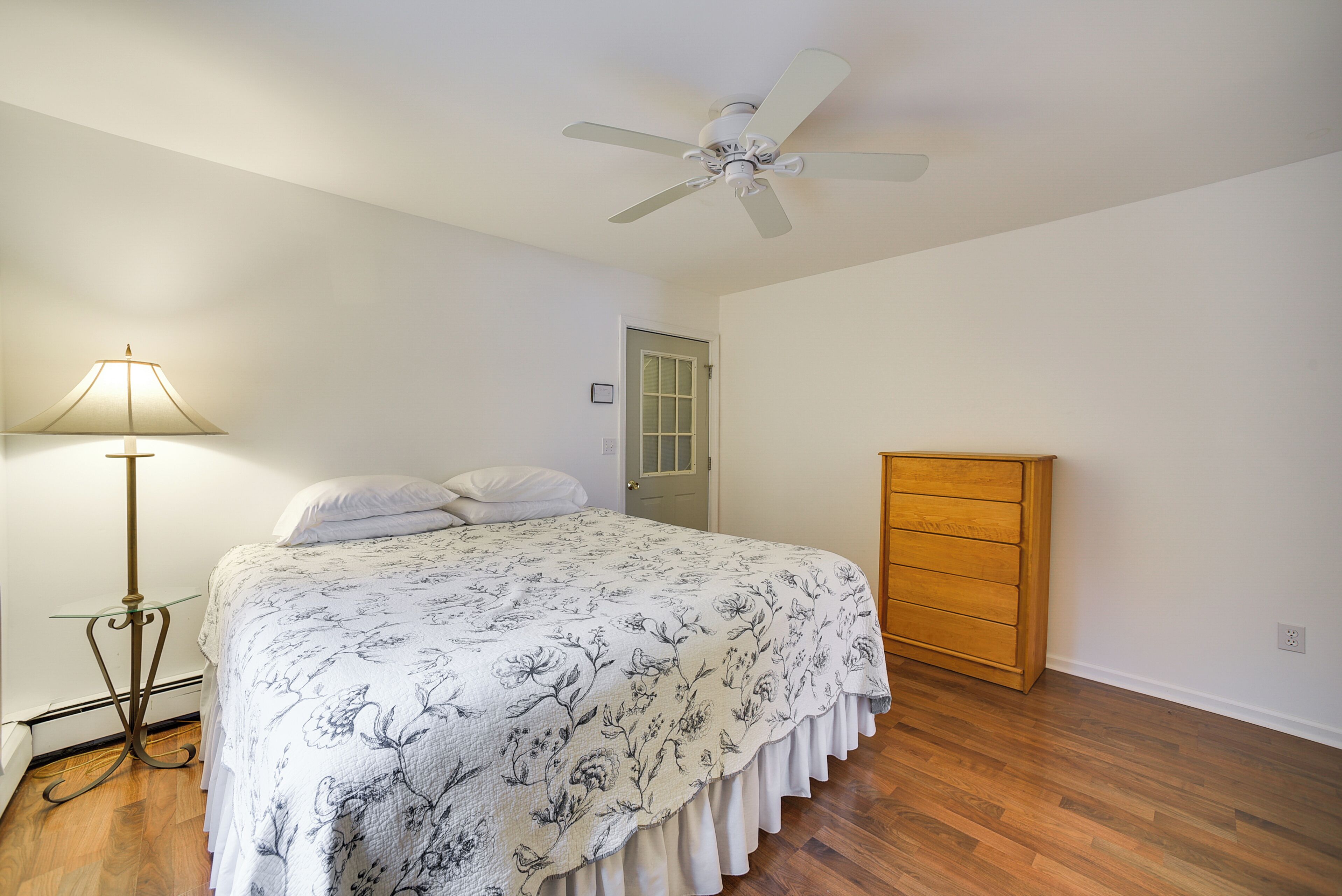 A bedroom with a bed, a bedside table, a lamp, a ceiling fan, a wooden dresser, and a door.