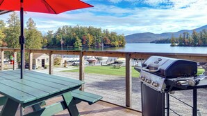 A balcony with a grill and picnic table, overlooking a lake and forest. - Lakefront Adirondacks Home: Fish, Hike, Ski! (Bolton Landing)