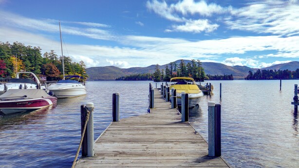 A wooden dock leading to boats on a calm lake with mountains in the background. - Lakefront Adirondacks Home: Fish, Hike, Ski! (Bolton Landing)