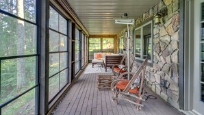 A covered porch with large windows, wicker chairs, a stone wall, and a view of the forest. - Thunder Lake Escape + Boat Dock! (Remer)