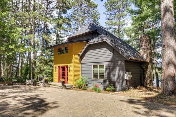 A two-story house with a yellow front and gray siding, surrounded by trees and a paved driveway. - Thunder Lake Escape + Boat Dock! (Remer)