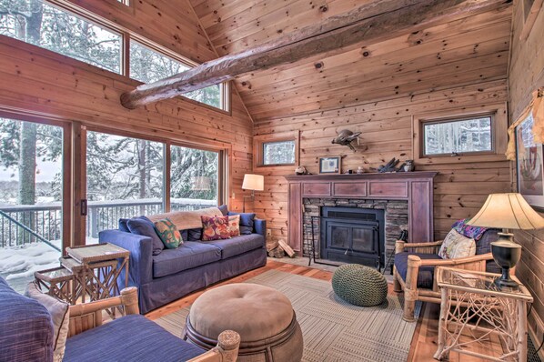 A cozy cabin interior with a fireplace, wooden walls, and a sofa by a large window overlooking a snowy landscape. - Thunder Lake Escape + Boat Dock! (Remer)