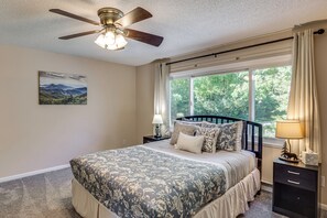 A bedroom with a bed, a nightstand, a lamp, a ceiling fan, a window with curtains, and a wall-mounted picture.