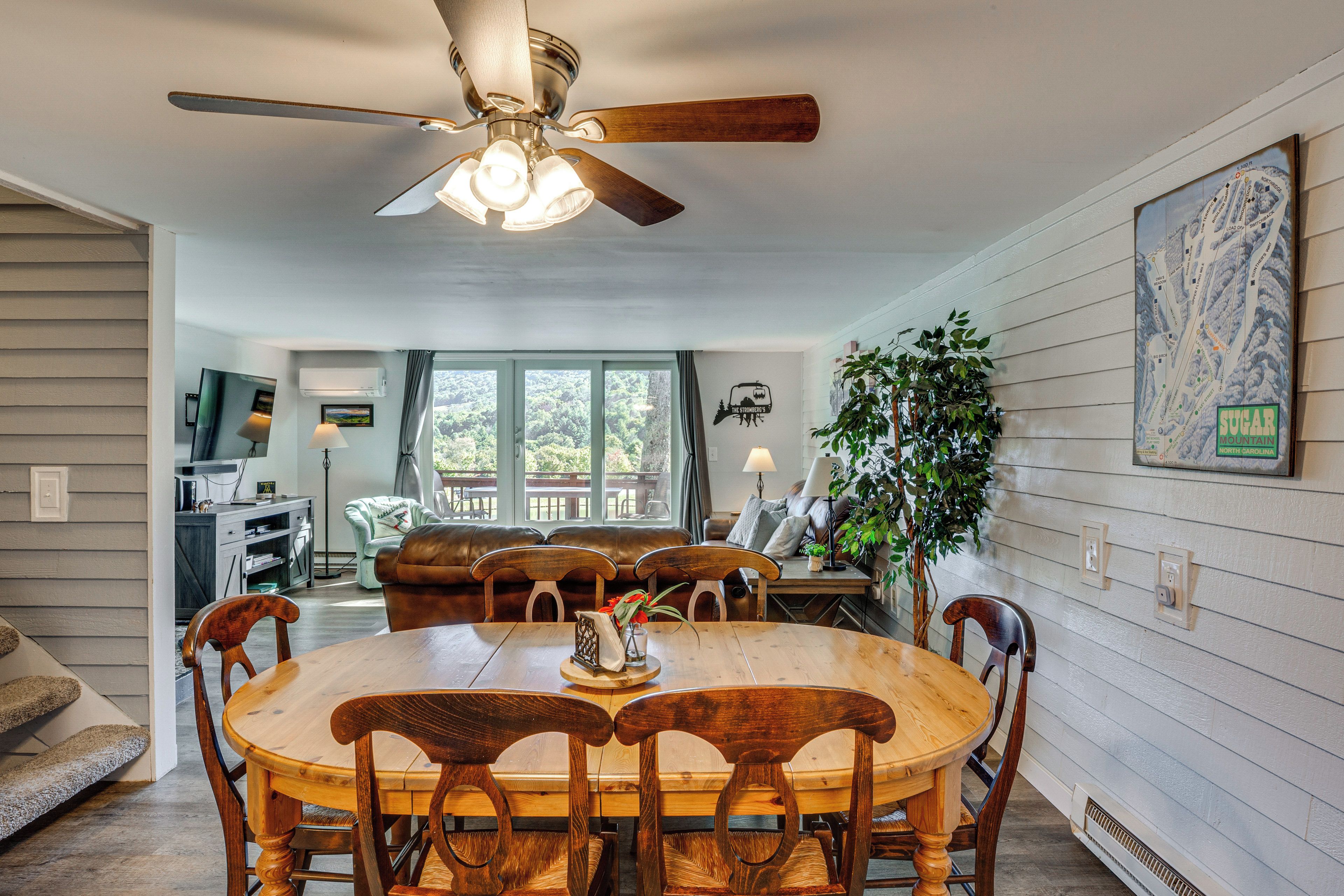 A dining area with a wooden table and chairs, a ceiling fan, and a view of the outdoors through large windows.