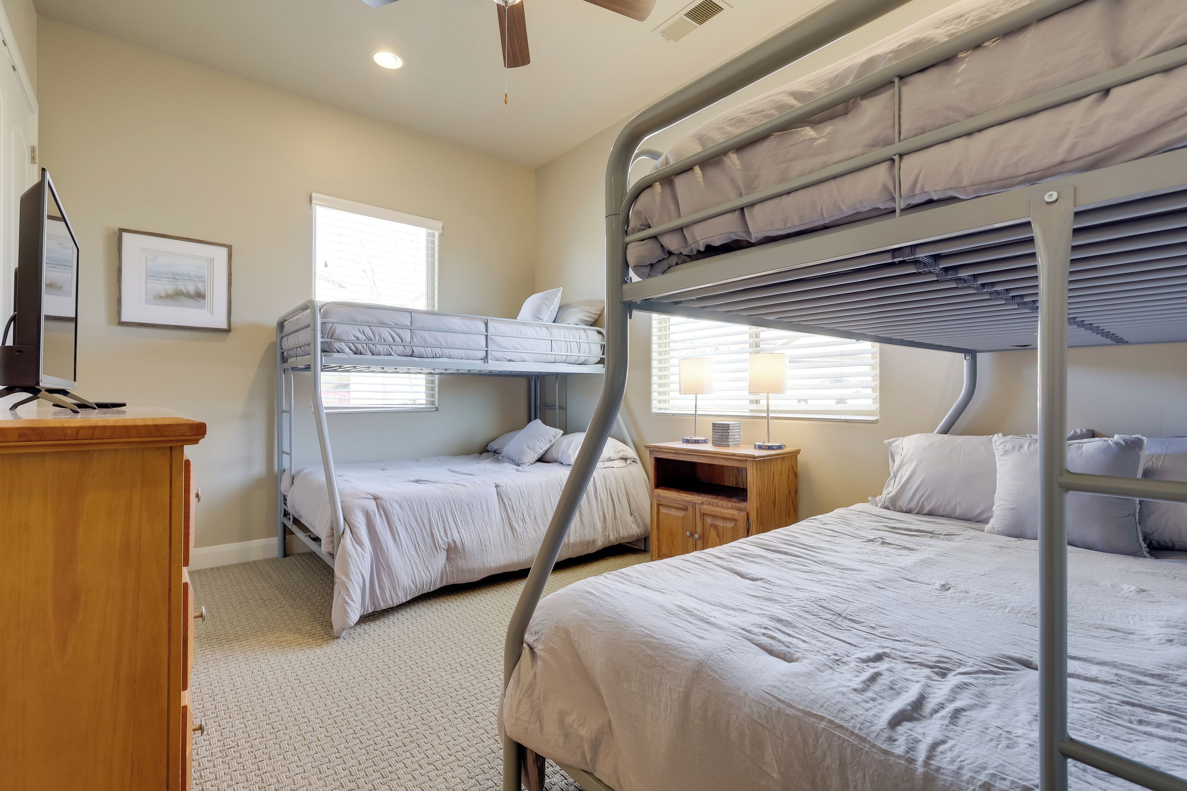 A dormitory room with bunk beds, a wooden dresser, a desk, and a window with blinds.