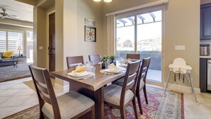 Un coin repas avec une table en bois, des chaises et une chaise haute. Une fenêtre offre une vue sur les maisons à l’extérieur.