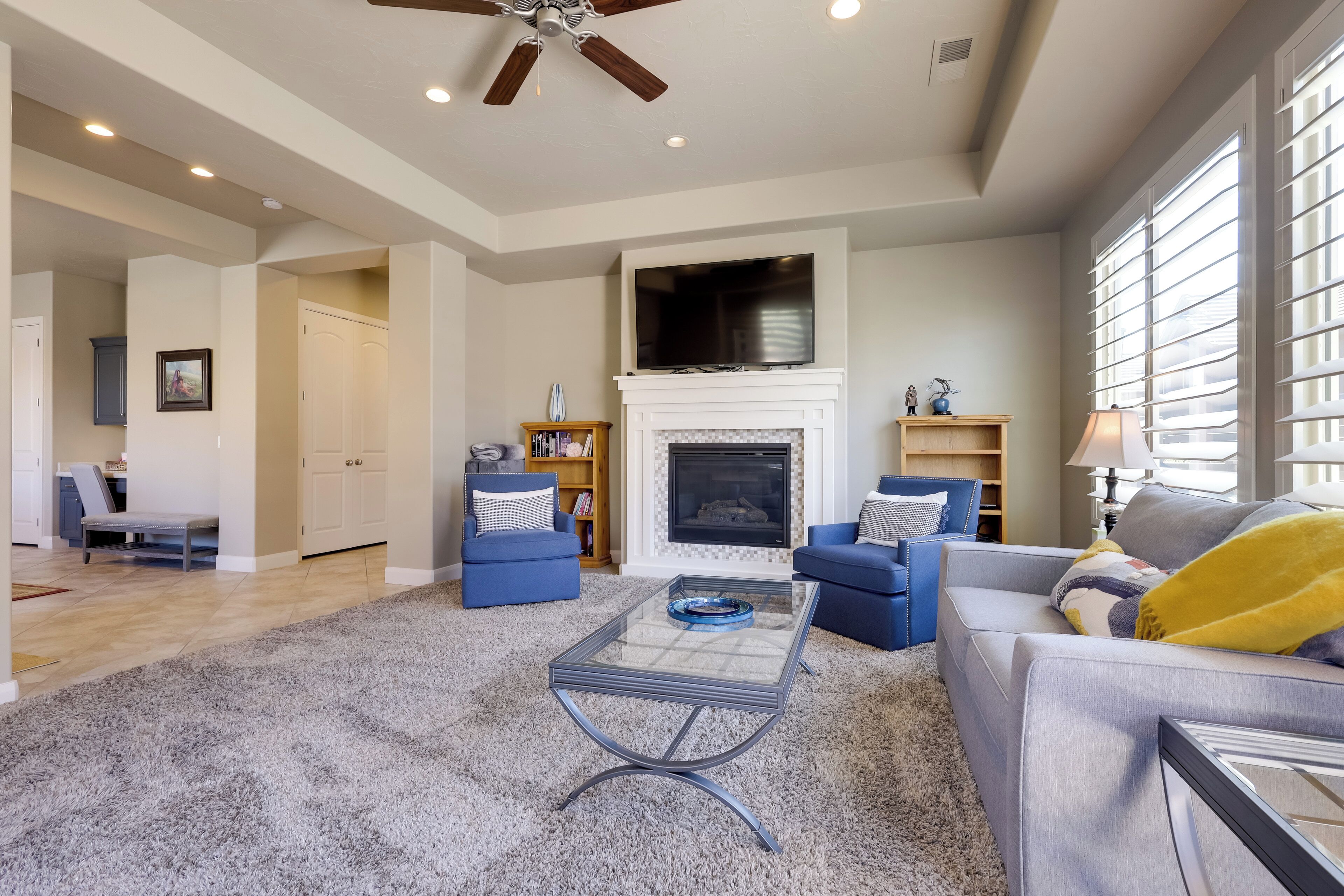 A spacious living room with a fireplace, a ceiling fan, and a large window with blinds.