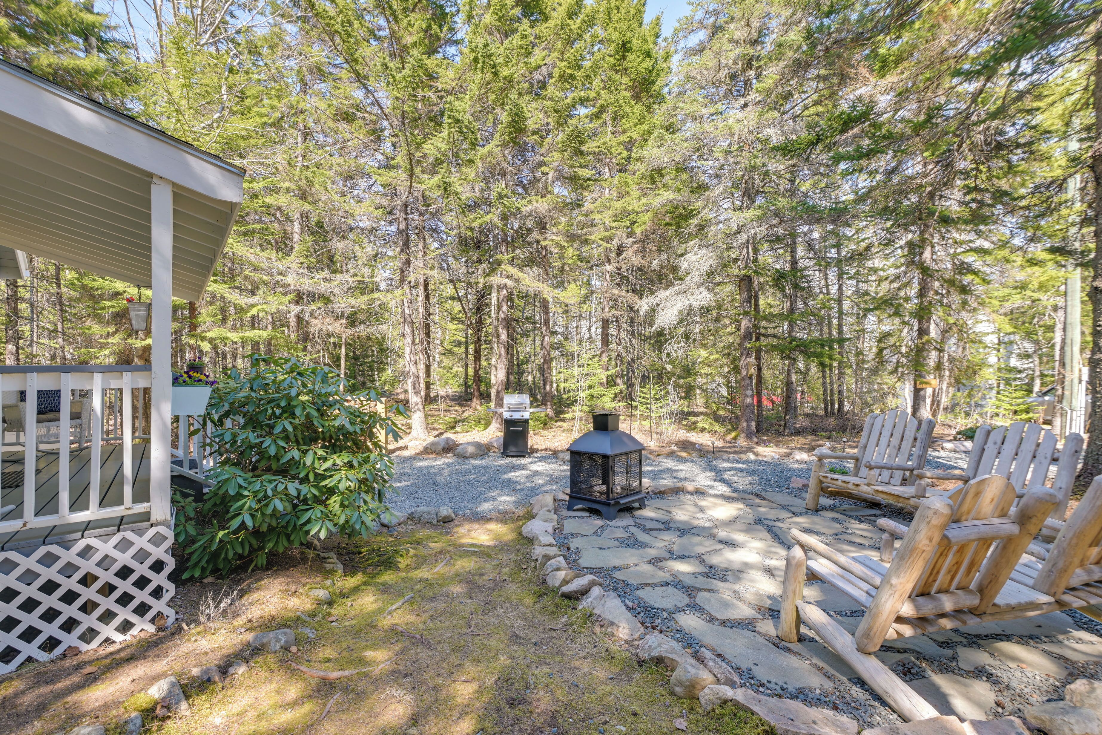 A cabin with a deck, surrounded by a forest. There are Adirondack chairs and a lantern.