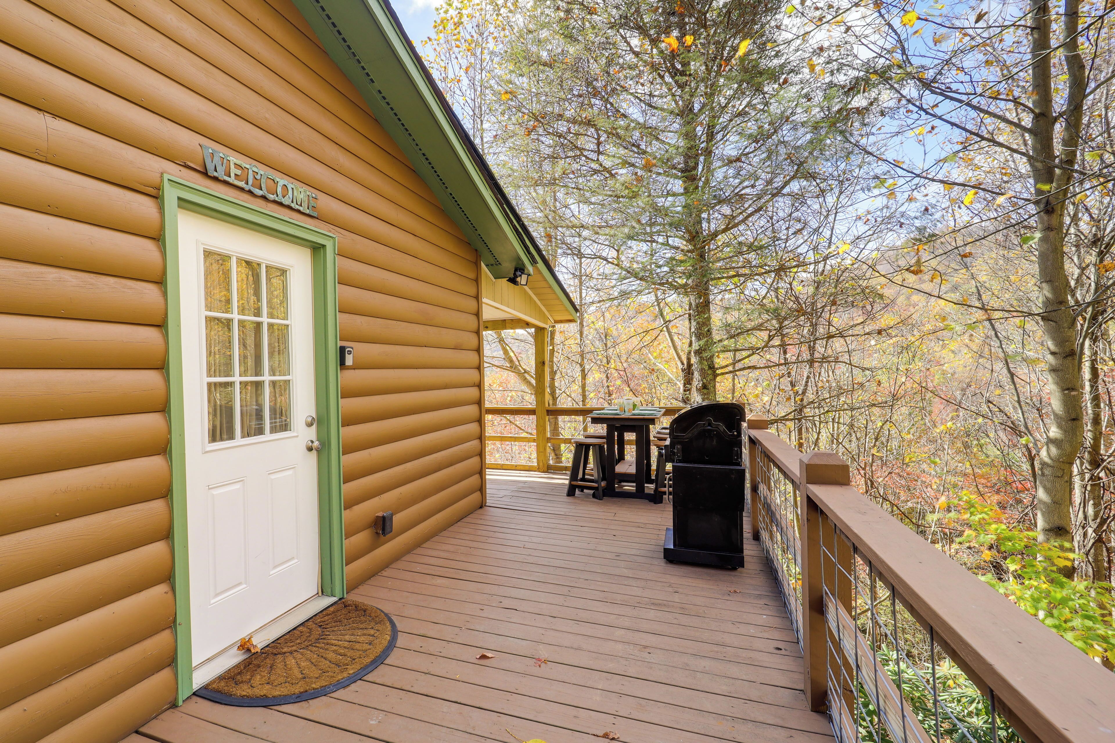 A wooden cabin with a 'WELCOME' sign, a deck with outdoor seating, and a view of trees and foliage.