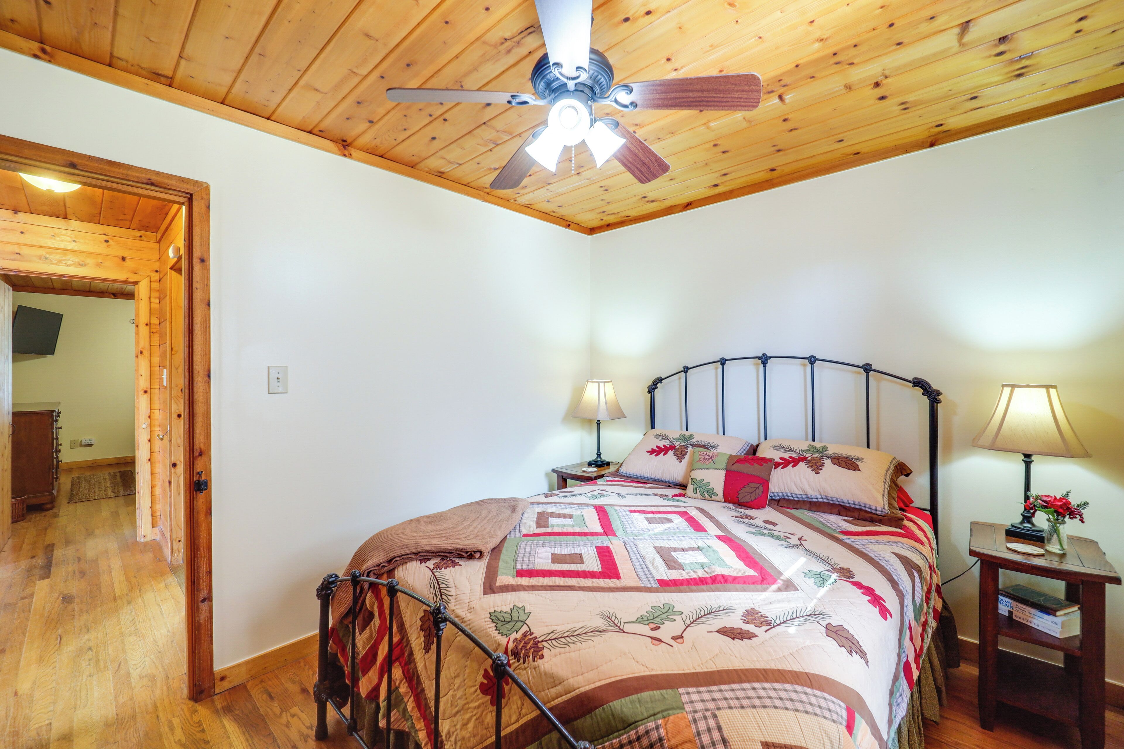 Un dormitorio con techo de madera, una cama con colcha, dos mesitas de noche con lámparas y una puerta que da a otra habitación.
