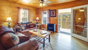 A cozy cabin interior with wooden walls, a ceiling fan, a fireplace, a sofa, a table, and a large window offering a view of the outdoors. - Secluded Lenoir Cabin: 4 Mi to Dtwn Blowing Rock (Lenoir)