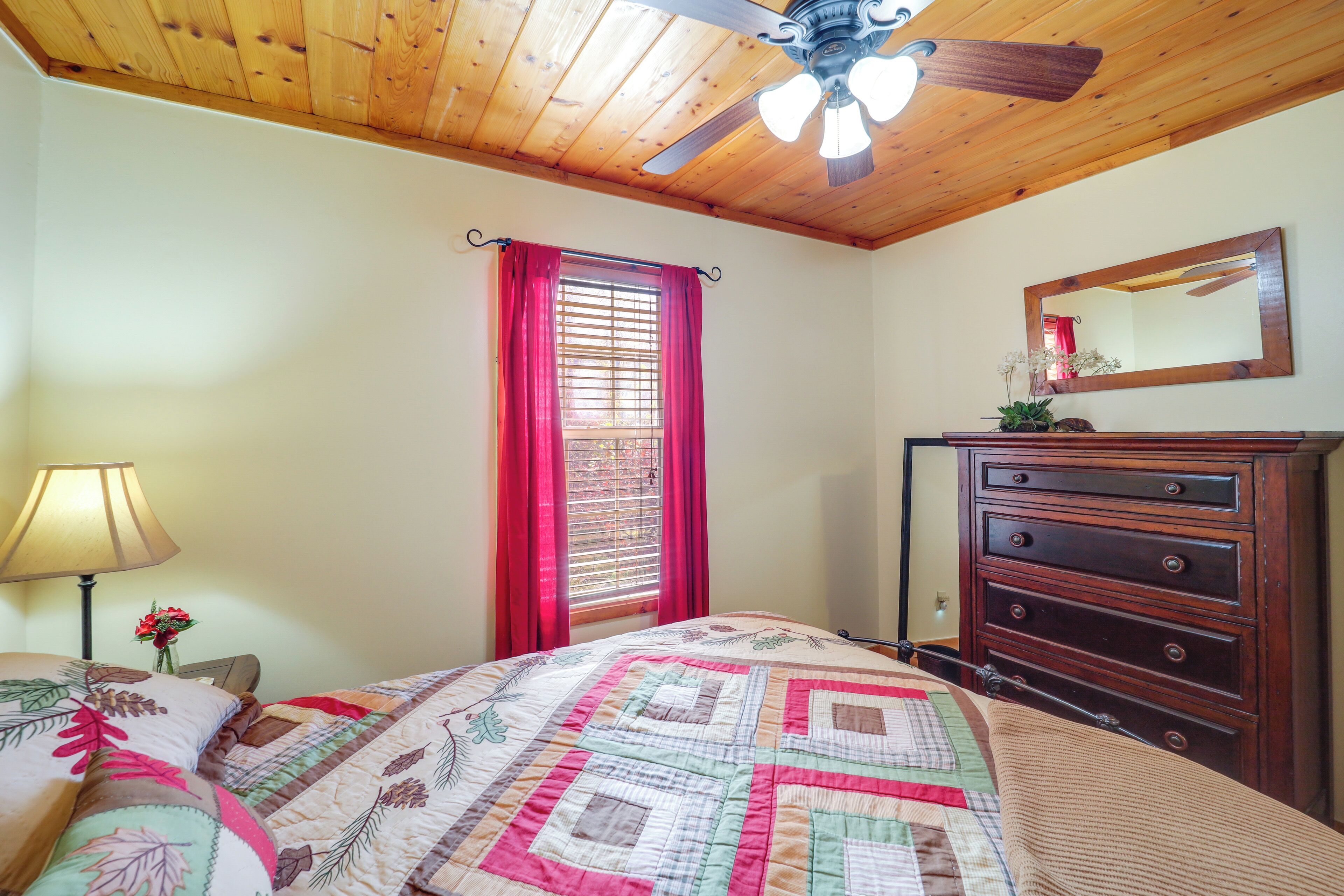 Une chambre avec un plafond en bois, un lit recouvert d ’une courtepointe, une commode, un miroir et une fenêtre avec des rideaux rouges.