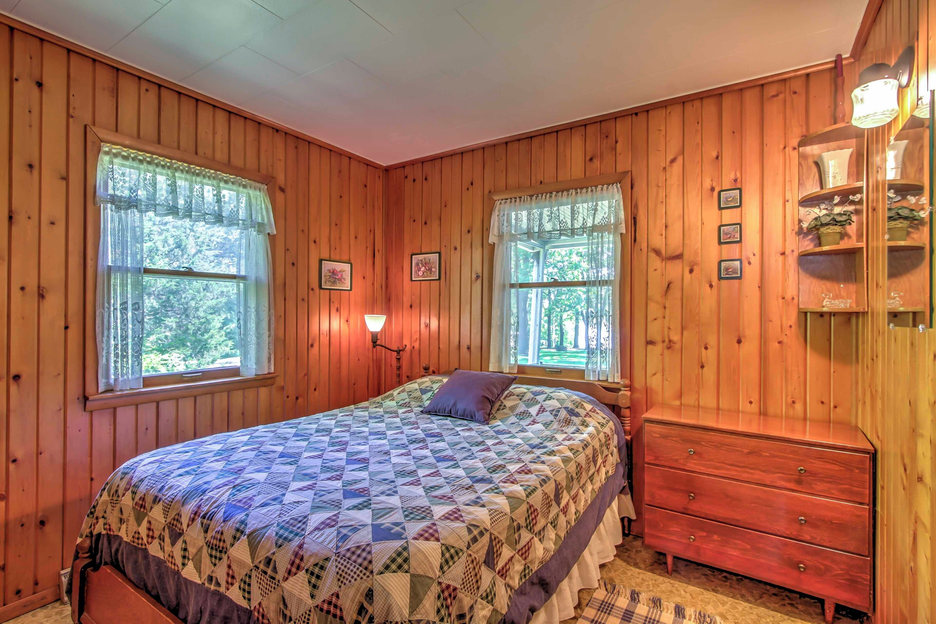 Un dormitorio con paneles de madera, una cama, dos ventanas con cortinas, un aparador y un estante de pared con adornos.