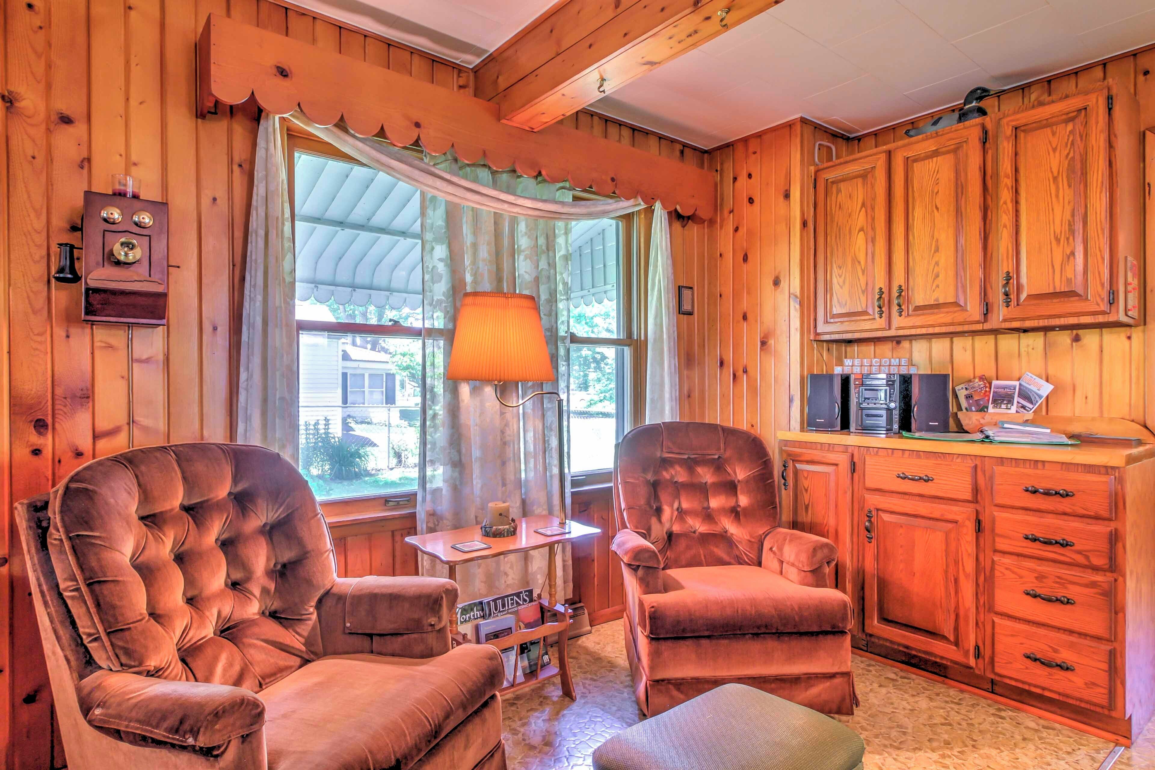 Habitación acogedora con paredes de madera y un ventanal con cortinas. Hay dos sillones cómodos, una mesita con lámpara y un mueble con electrodomésticos de cocina.