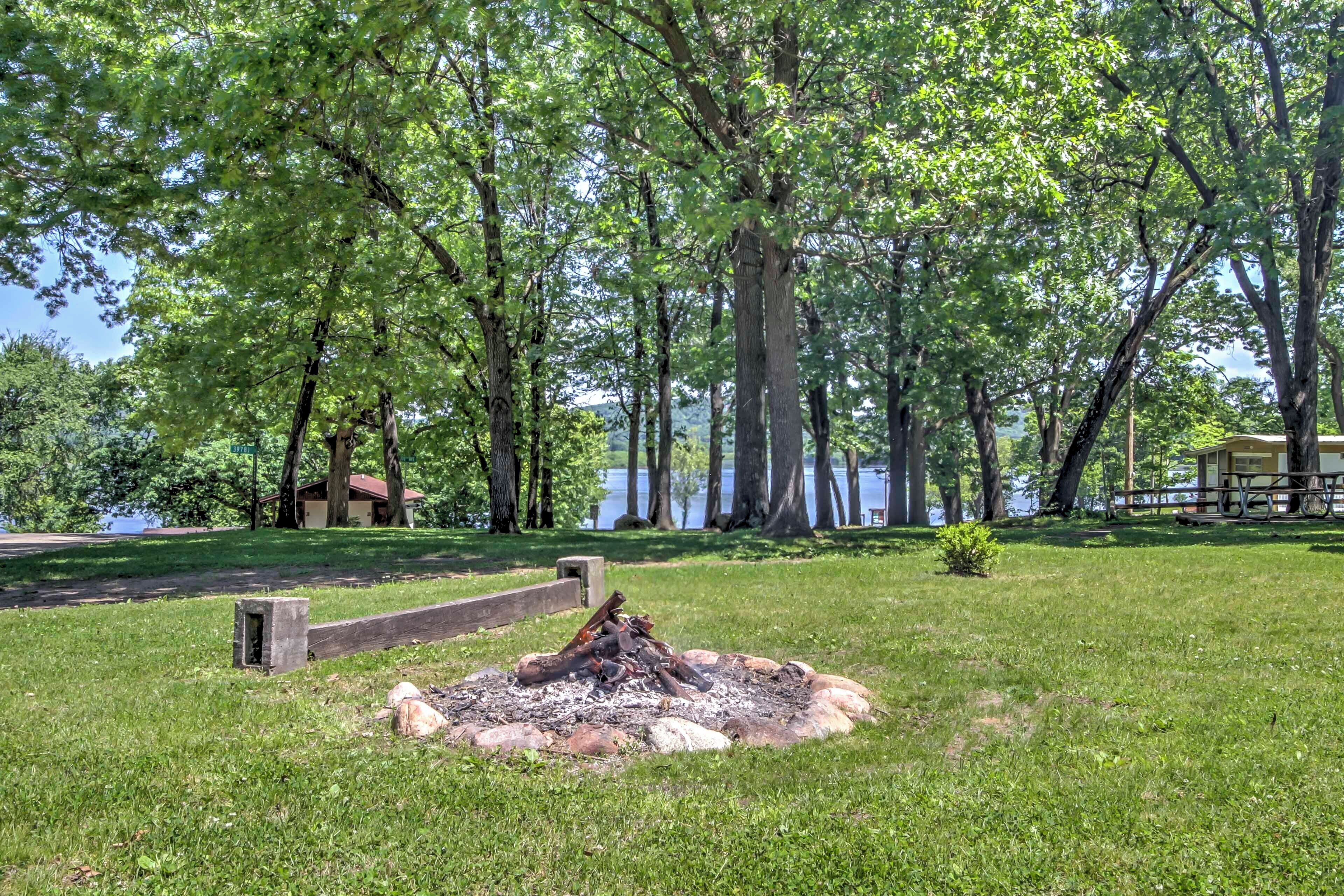 Een grasrijke plek met een vuurkuil, omringd door rotsen, bomen en een waterpartij op de achtergrond.