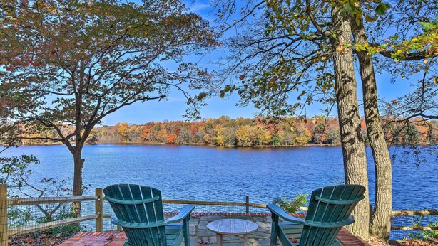 A lakeside seating area with two Adirondack chairs and a small table, surrounded by trees and a view of the lake.