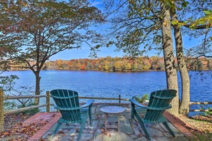 A lakeside seating area with two Adirondack chairs and a small table, surrounded by trees and a view of the lake. - Vibrant Milford Home w/ Boat Dock & Patio! (Milford)