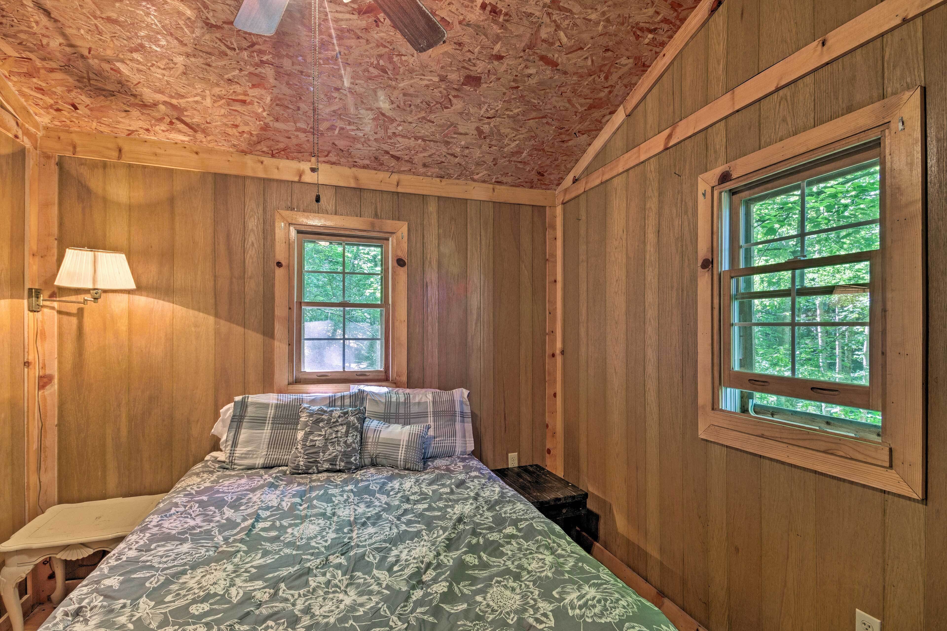 A wooden-paneled bedroom with a bed, two windows, and a ceiling fan.