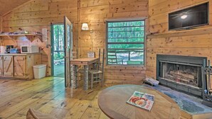 A wooden cabin interior with a fireplace, a television mounted above it, a dining table with a magazine, and a kitchen area with a microwave. - Pisgah Forest 'the Oak' Cabin w/ Deck by Creek (Barnardsville)