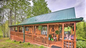 A wooden cabin with a green roof, a porch with railings, and various outdoor items like a flag and a barrel. - Wellington Cabin Escape w/ Wraparound Deck! (Wellington)