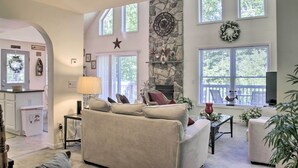 A spacious living room with a stone fireplace, a sofa, and a television. There are large windows and a kitchen visible in the background. - Spacious Home w/ Deck, Grill & Delaware River View (Callicoon)