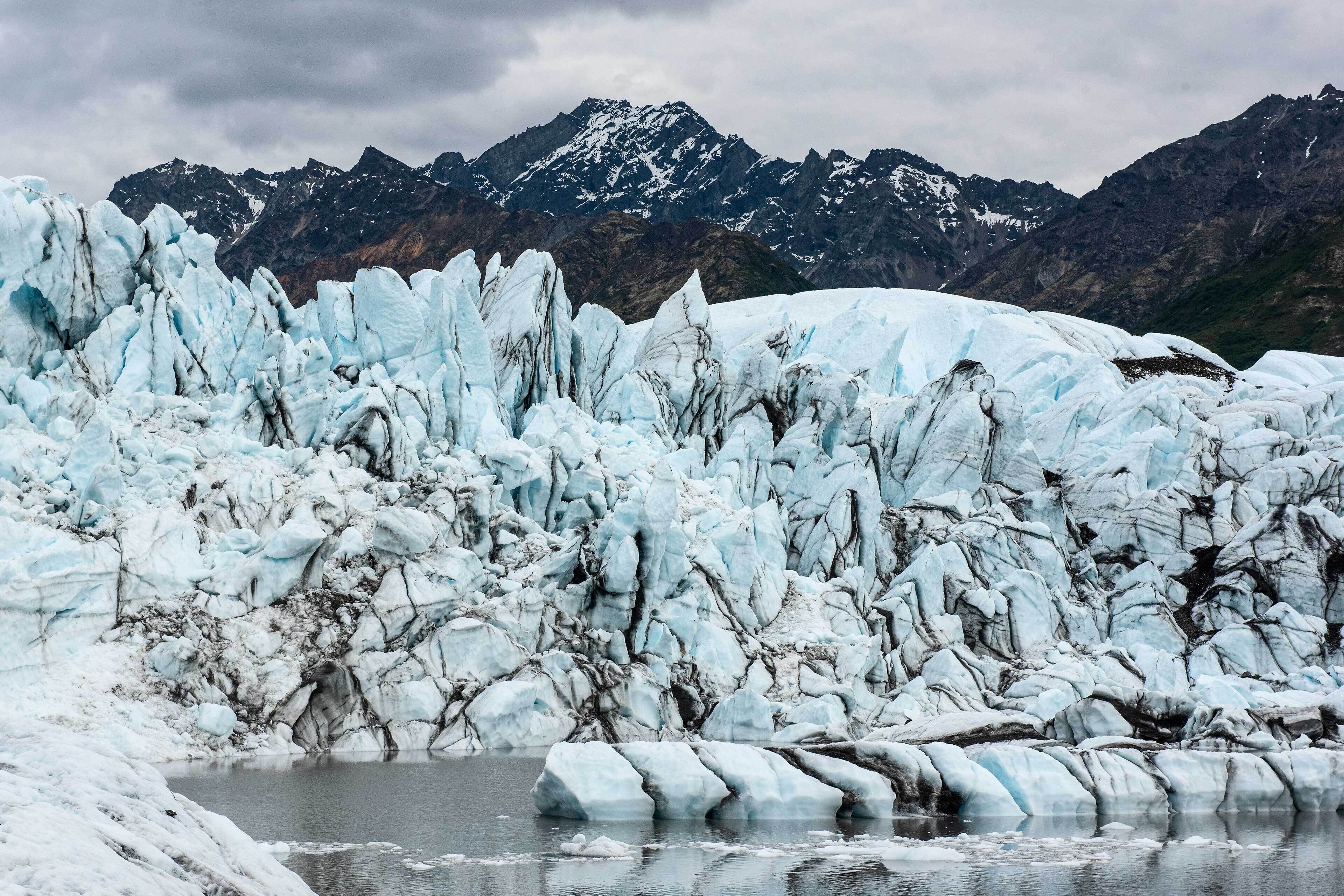 A glacier with jagged ice formations and a body of water in the foreground, with mountains in the background.