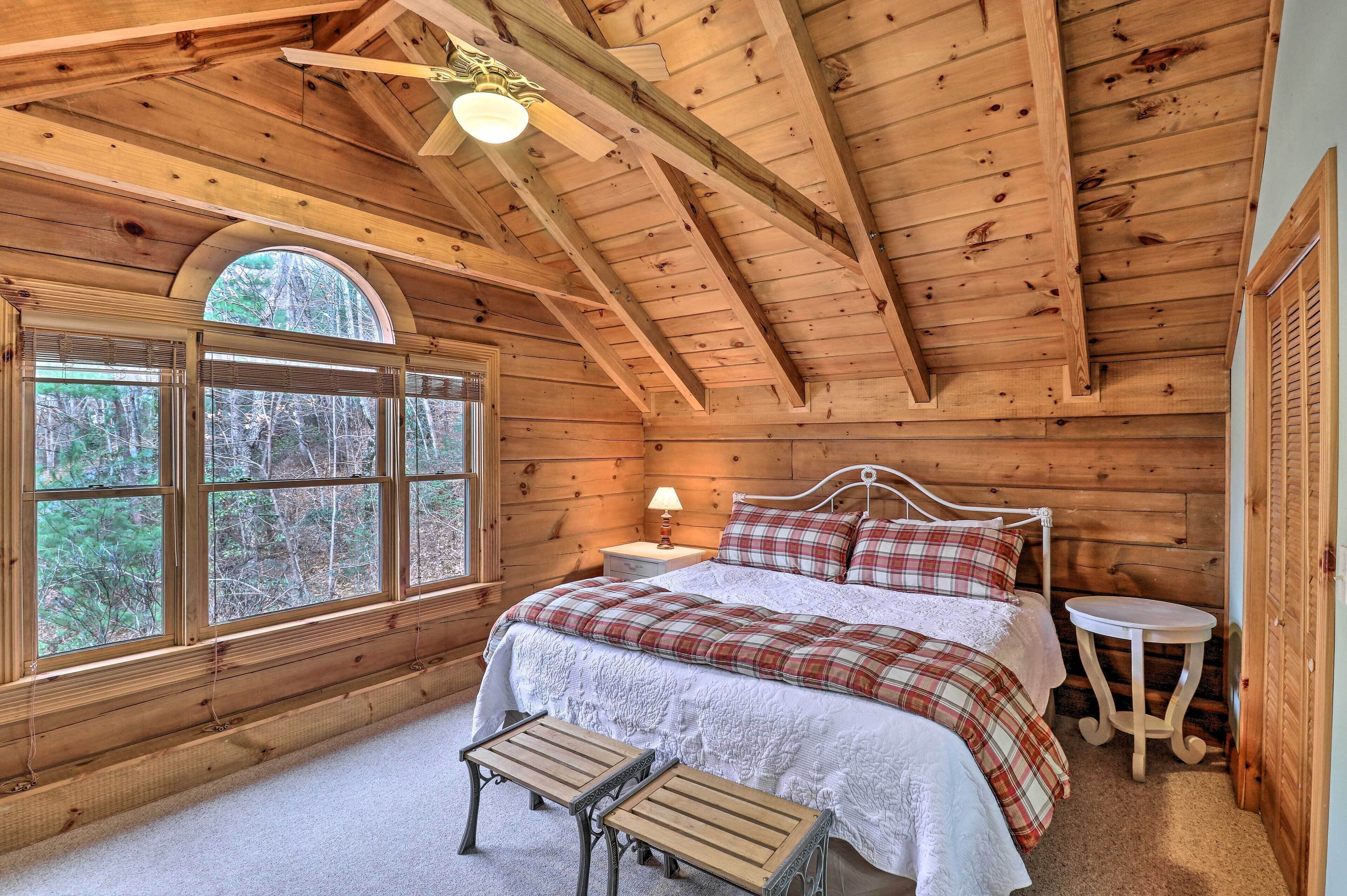 Un dormitorio de cabaña de madera con un ventanal, una cama con ropa de cama escocesa, una mesita de noche con una lámpara y una pequeña mesa redonda.