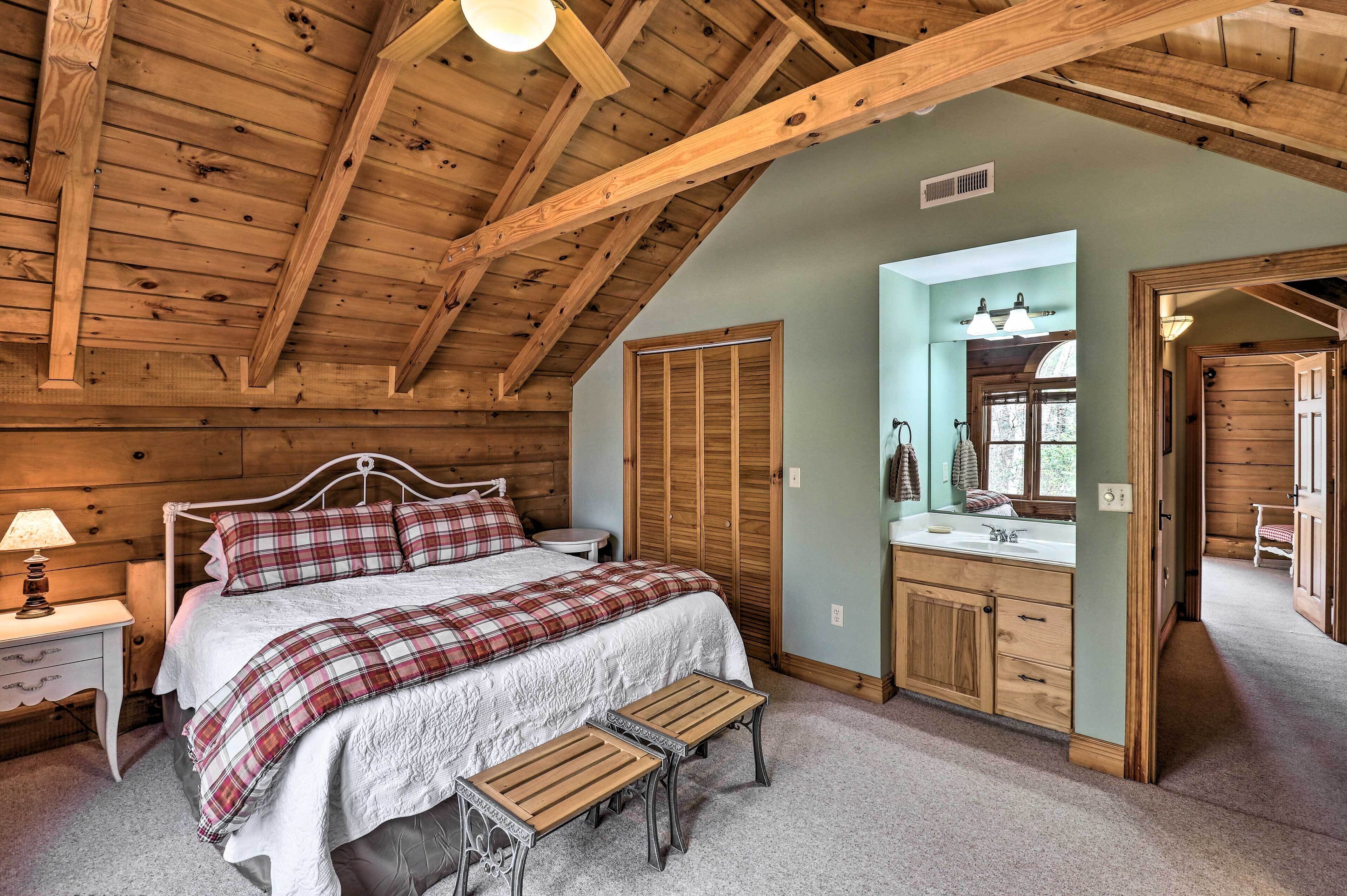 Un dormitorio con techo de madera, una cama con manta escocesa, una mesita de noche con lámpara, un banco de madera y un baño con espejo y lavamanos.