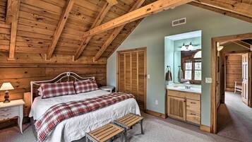 Un dormitorio con techo de madera, una cama con manta escocesa, una mesita de noche con lámpara, un banco de madera y un baño con espejo y lavamanos.