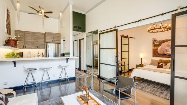 A modern hotel room with a kitchen area, a dining table, bar stools, a bed with a large window, and a chandelier.