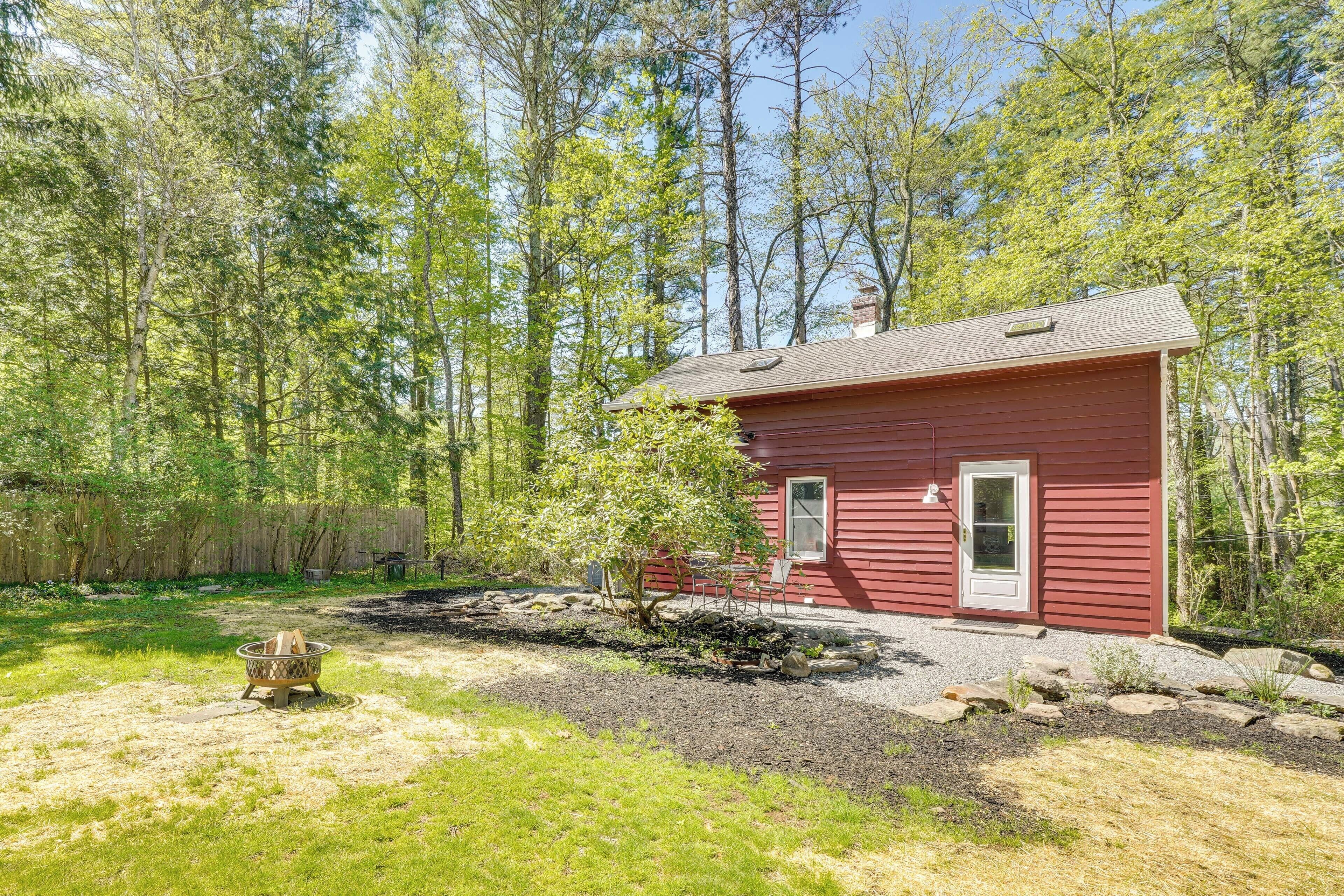 A red cabin with a door and window, surrounded by a forest and a fire pit in the yard.