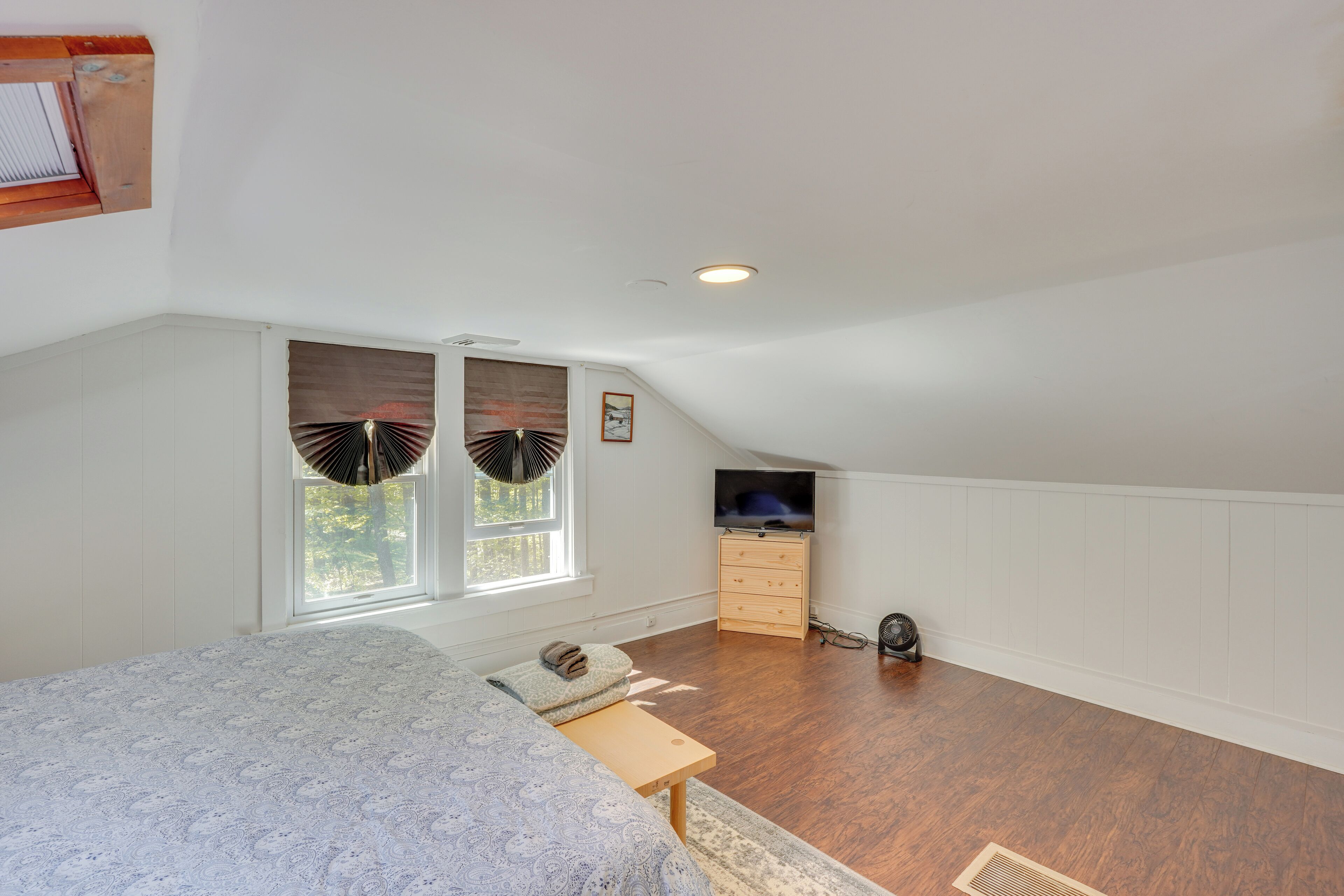 A bedroom with a sloped ceiling, a bed, a dresser, a TV, and a window with blinds.