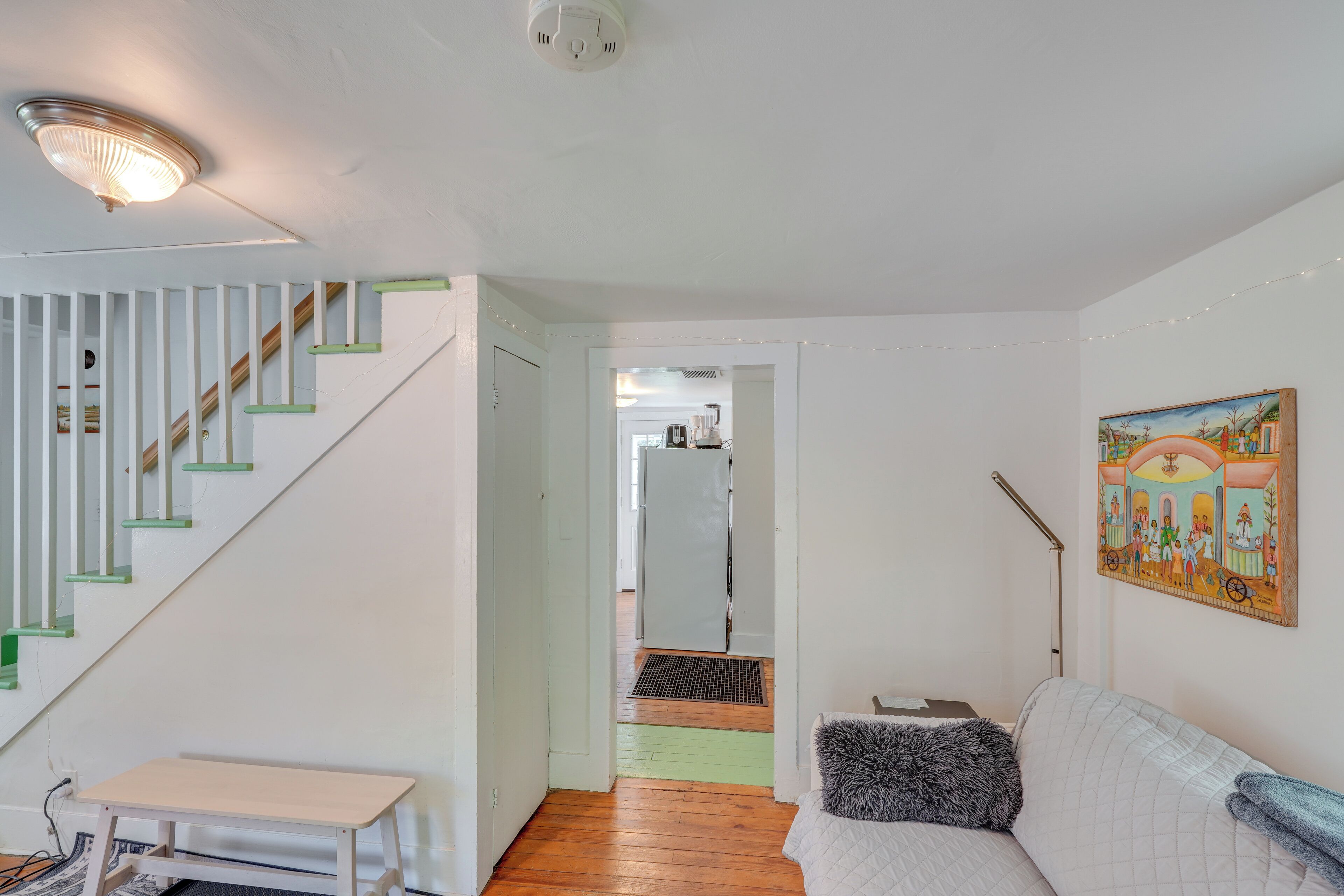 A room with a staircase, a bed, a small table, a painting, and a door leading to another room.