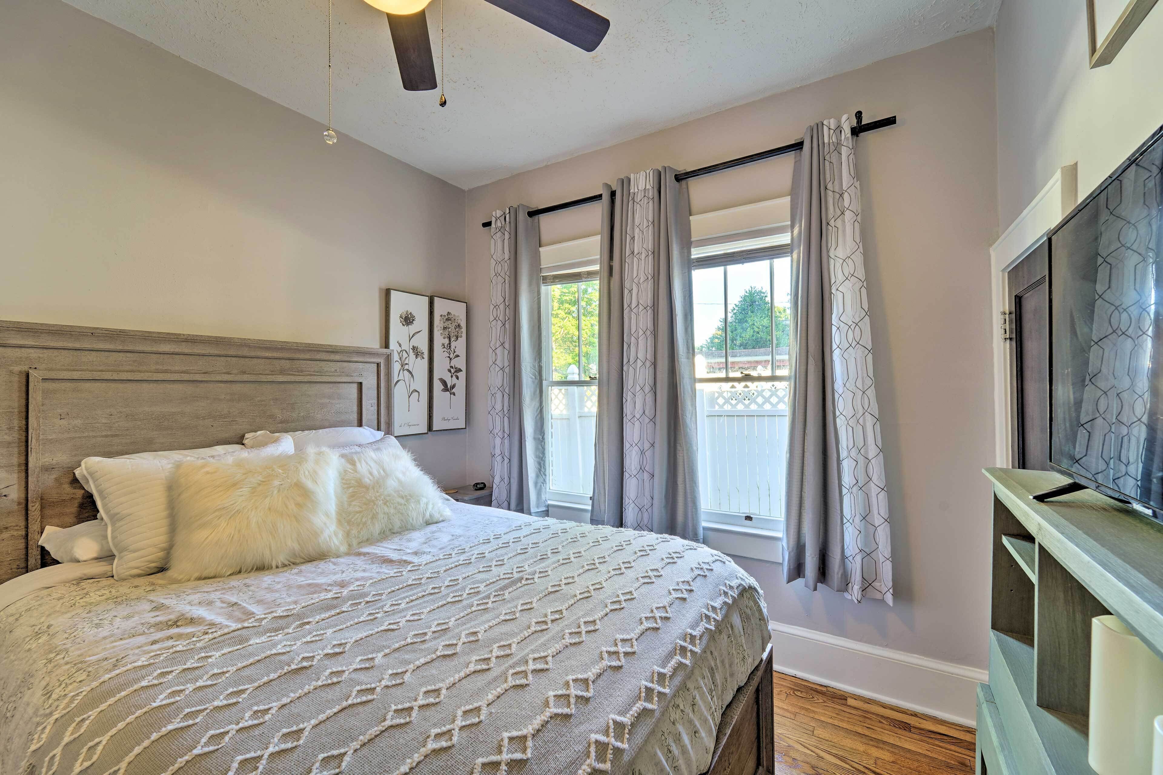 A bedroom with a wooden headboard, a bed with a patterned bedspread, a television on a wooden stand, and a window with curtains.