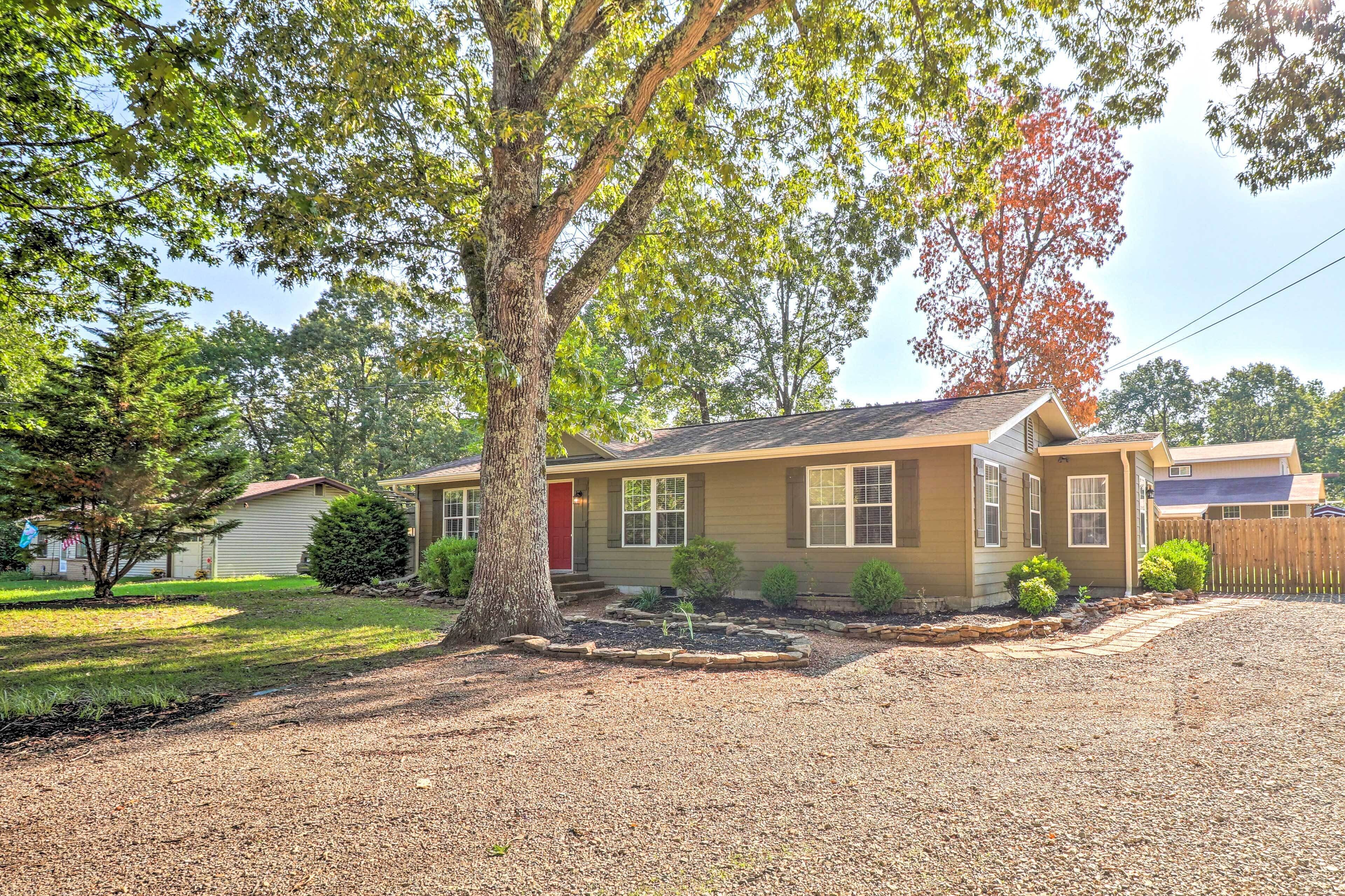 A single-story house with a red door, a front yard with a tree, and a gravel driveway.