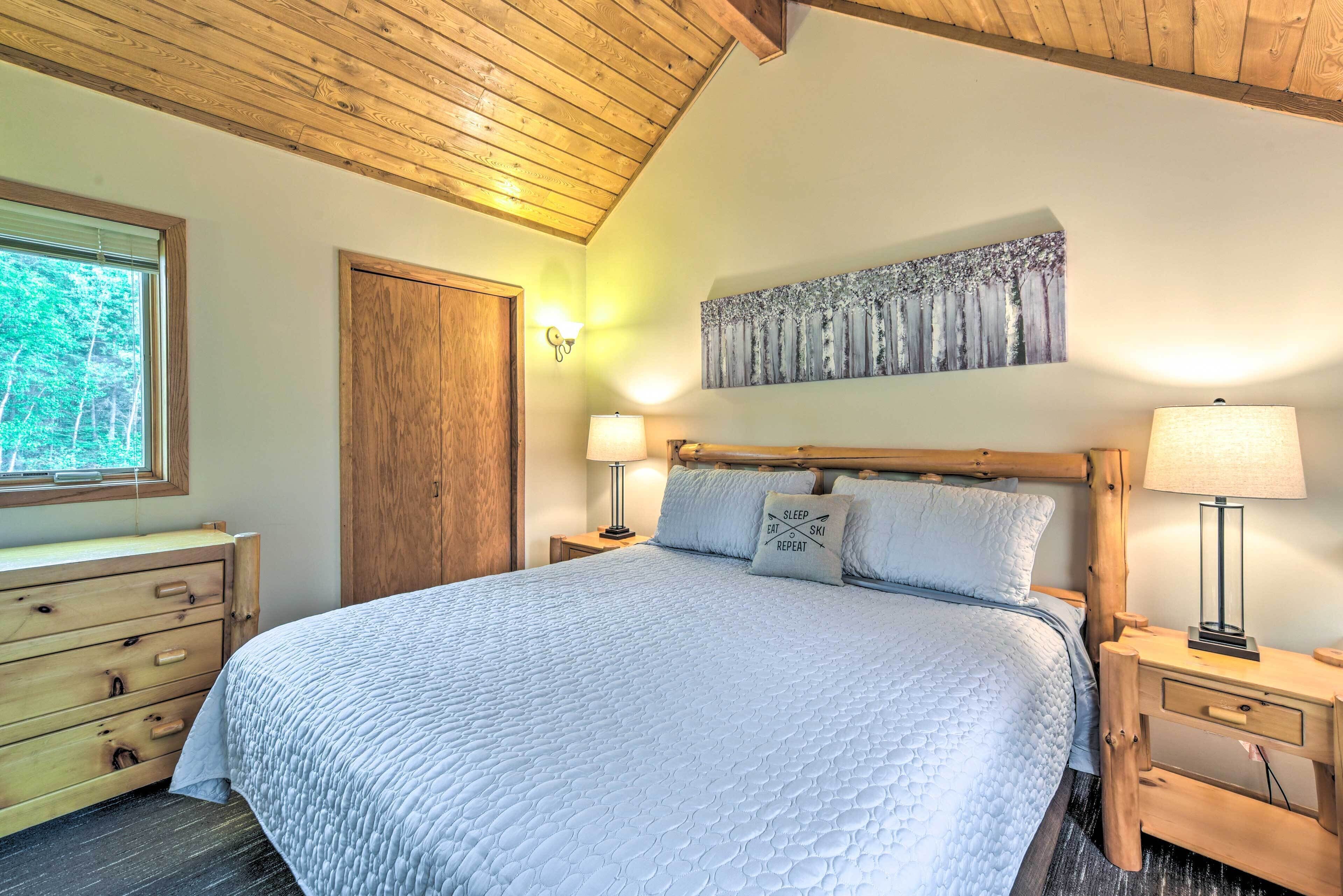 Une chambre à coucher avec un grand lit, une tête de lit en bois, deux tables de chevet avec des lampes, une commode en bois et une fenêtre avec des stores.