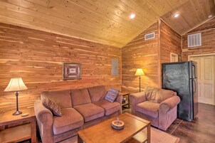 A cozy cabin interior with wooden walls, a sofa, a chair, a table, a refrigerator, and two lamps.
