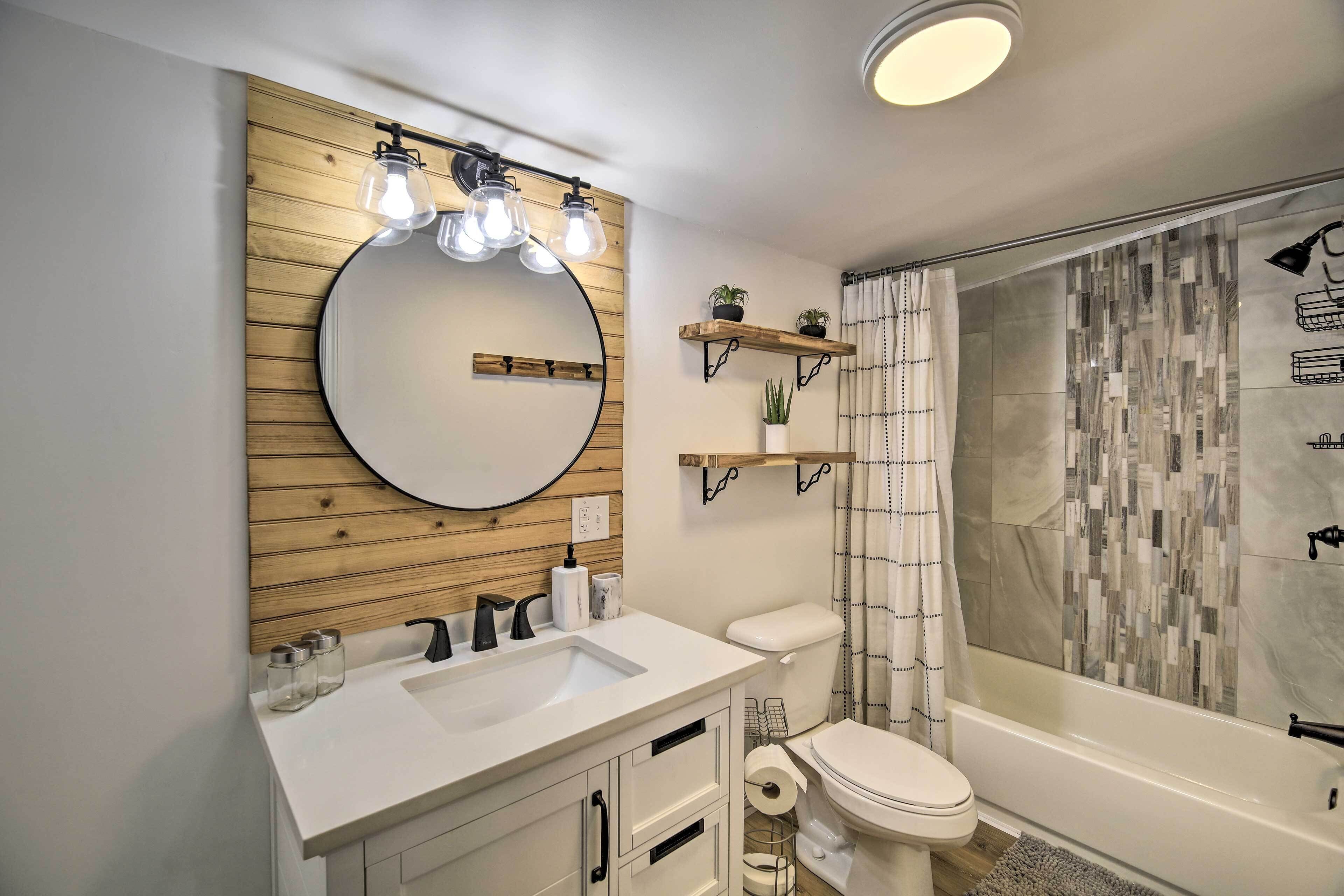 A modern bathroom with a wooden vanity, a round mirror, a toilet, and a shower with a mosaic tile wall.
