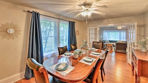 A dining room with a wooden table set for six, surrounded by chairs. There is a ceiling fan above and a large window with curtains.