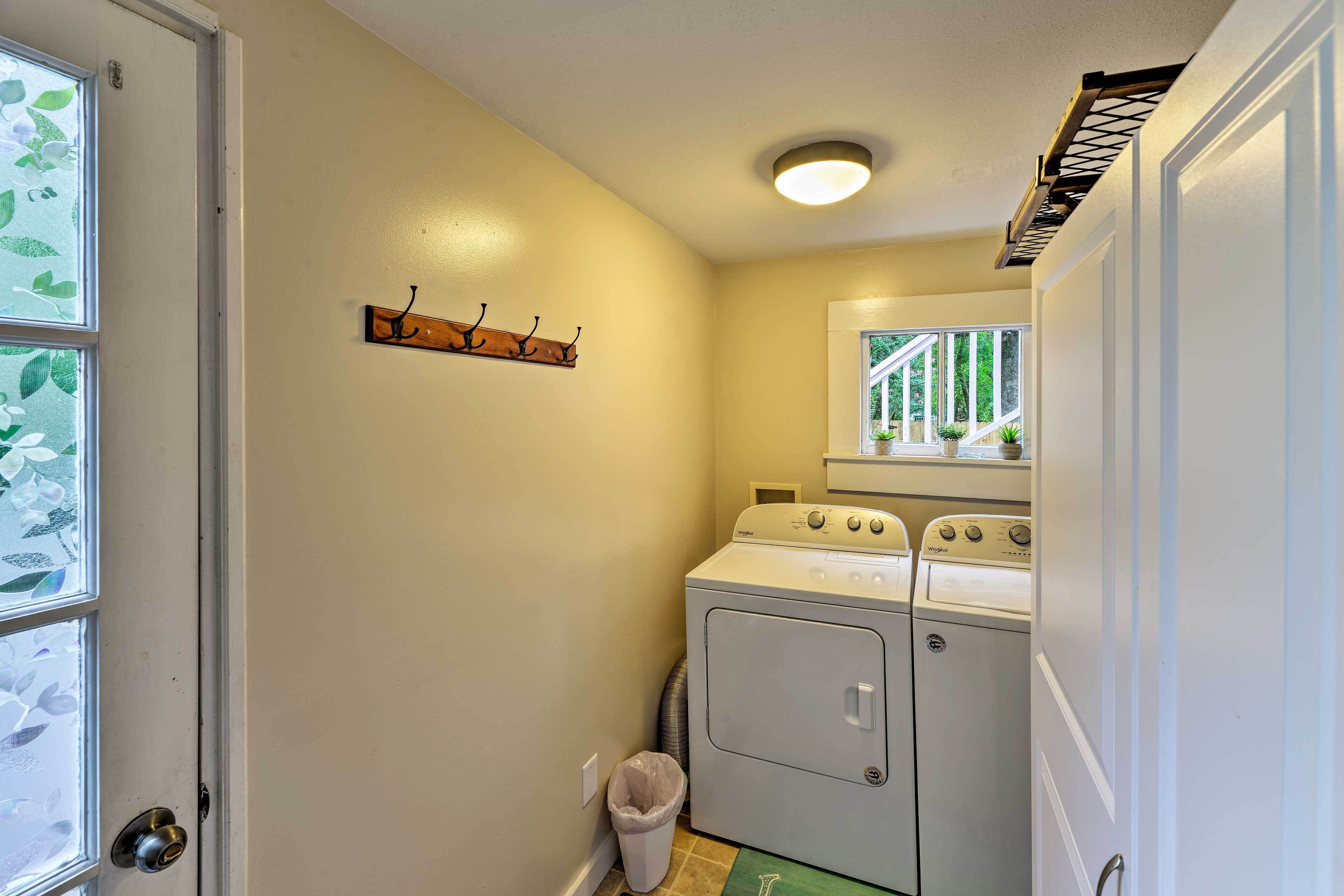 A laundry room with a washer and dryer, a trash can, and a window with a view of greenery.