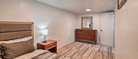 A bedroom with a wooden headboard, a bed with patterned bedding, a nightstand with a lamp, a dresser with drawers, and a door.