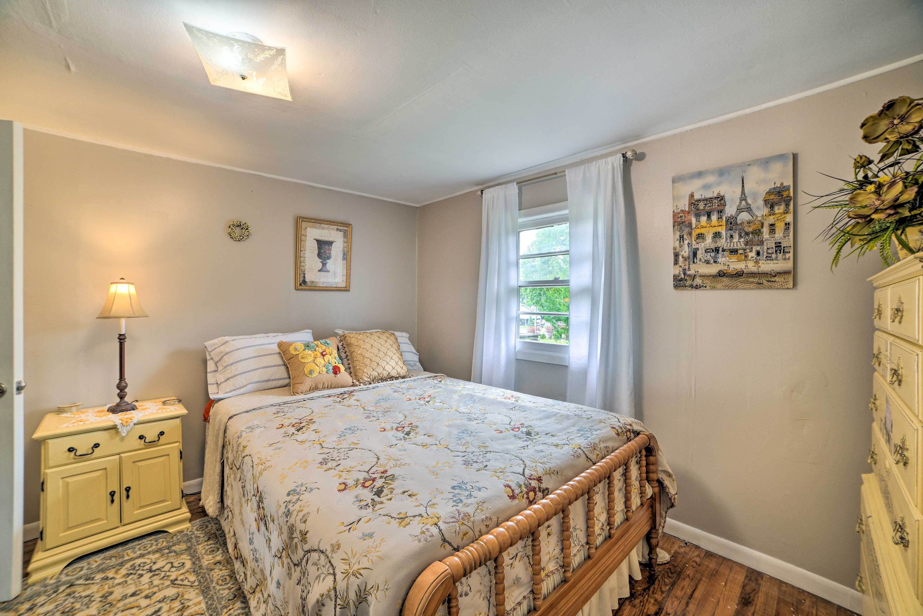 A bedroom with a floral bedspread, a bedside lamp, a dresser, and a painting of a cityscape.